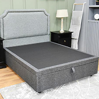 A gray upholstered storage bed frame with a dark gray mattress, shown in a bedroom setting with a nightstand, lamp, and wall panels.