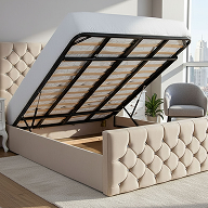 A beige upholstered bed with a tufted headboard and footboard, shown in a raised position to reveal the black metal frame and storage compartment. The bed is photographed in a bright bedroom setting with a gray armchair and a window in the background.